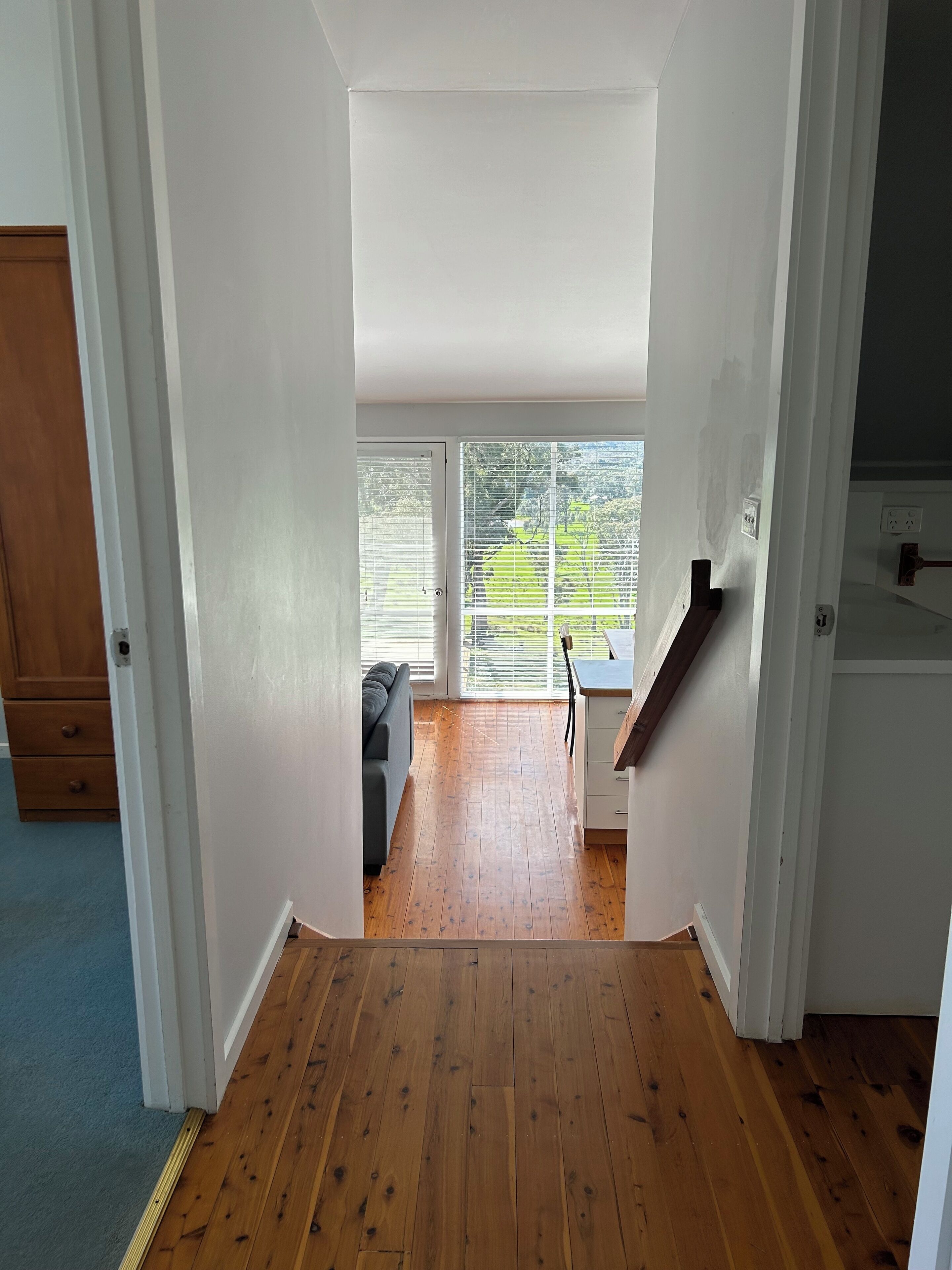 A hallway with wooden flooring leading to a room with a large window, a sofa, and a desk.