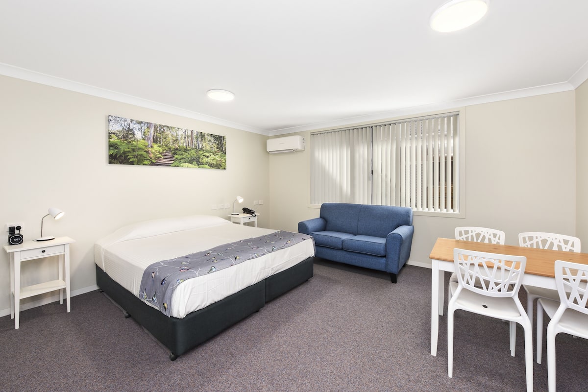 A hotel room with a bed, a blue sofa, a dining table with white chairs, a small desk with a lamp, and a picture on the wall.