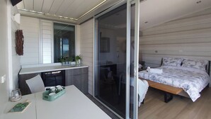 Un dormitorio moderno con una cama grande, un ventanal corredero a un balcón, una pequeña cocina y una mesa de comedor con una bandeja verde.