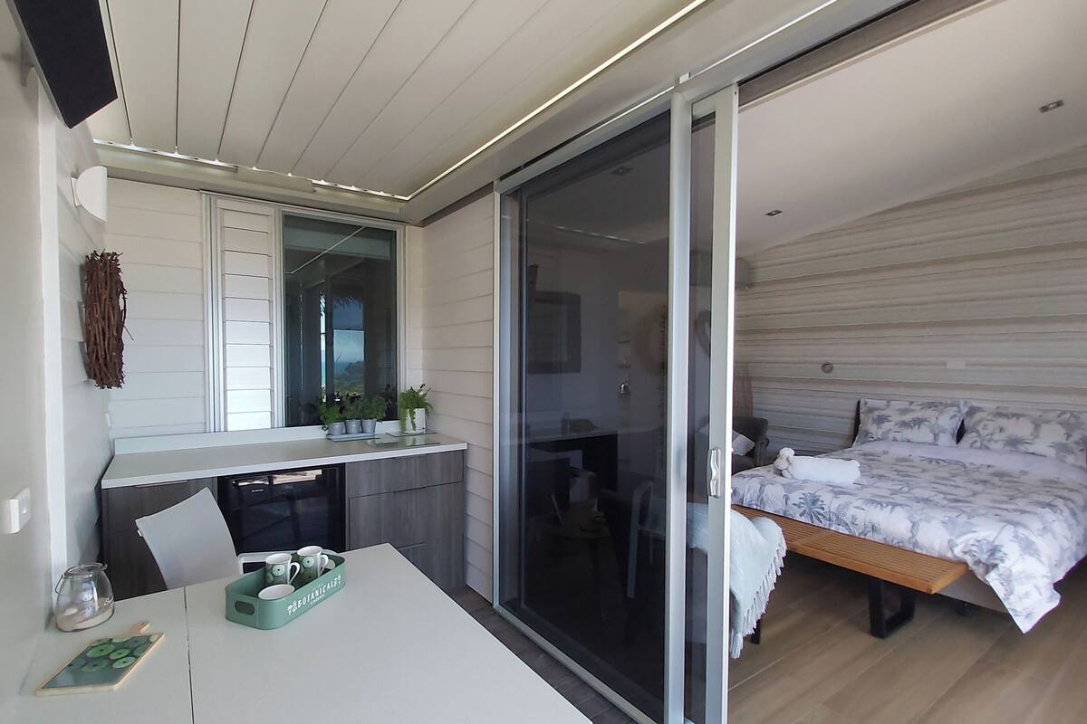 A modern bedroom with a large bed, a sliding glass door to a balcony, a small kitchen area, and a dining table with a green tray.