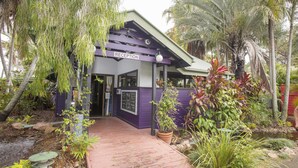 Zona d'entrada amb façana morada, passatge de fusta i plantes tropicals.