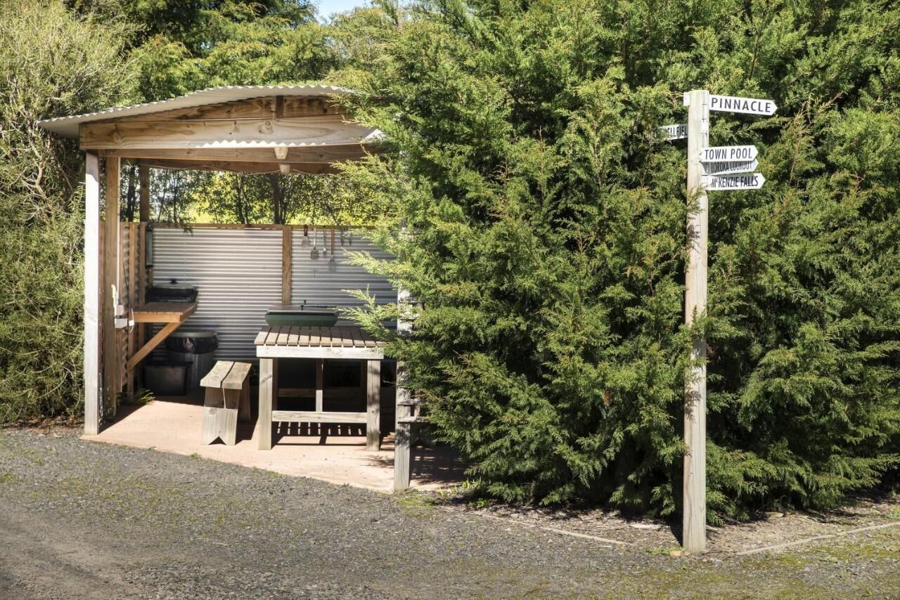 Un abri rustique en bois, doté d’un banc et d’une table, est entouré d’arbres à feuilles persistantes denses. Un panneau indicateur signale les lieux à proximité.