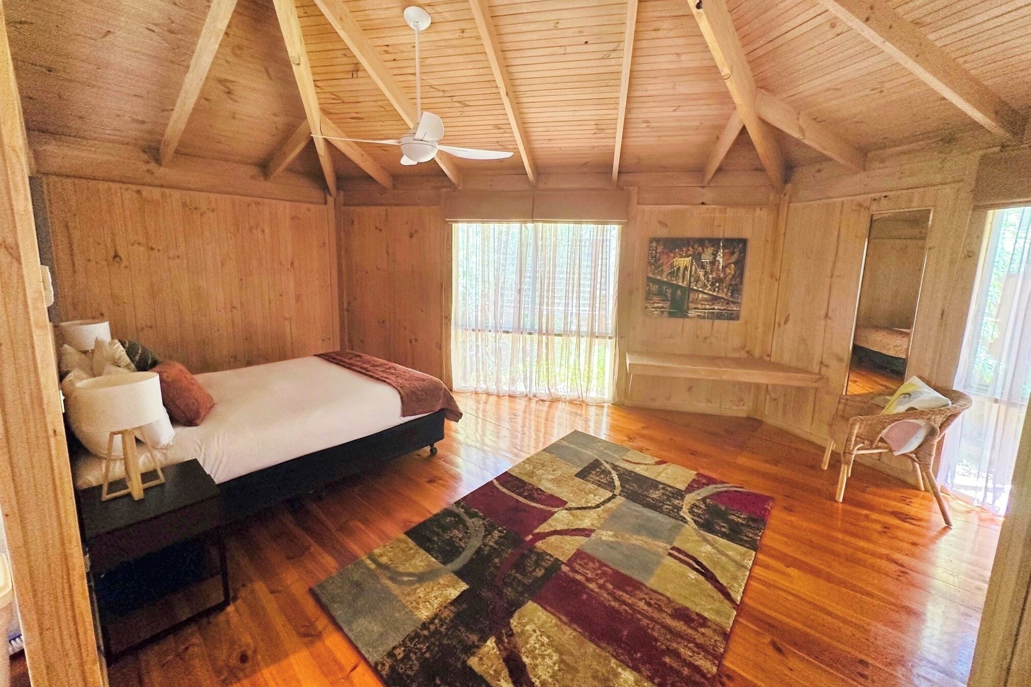 Un dormitorio de cabaña de madera con una cama, un ventilador de techo, una ventana con cortinas translúcidas, un cuadro en la pared y una silla pequeña.