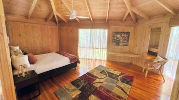 Un dormitorio de cabaña de madera con una cama, un ventilador de techo, una ventana con cortinas translúcidas, un cuadro en la pared y una silla pequeña.