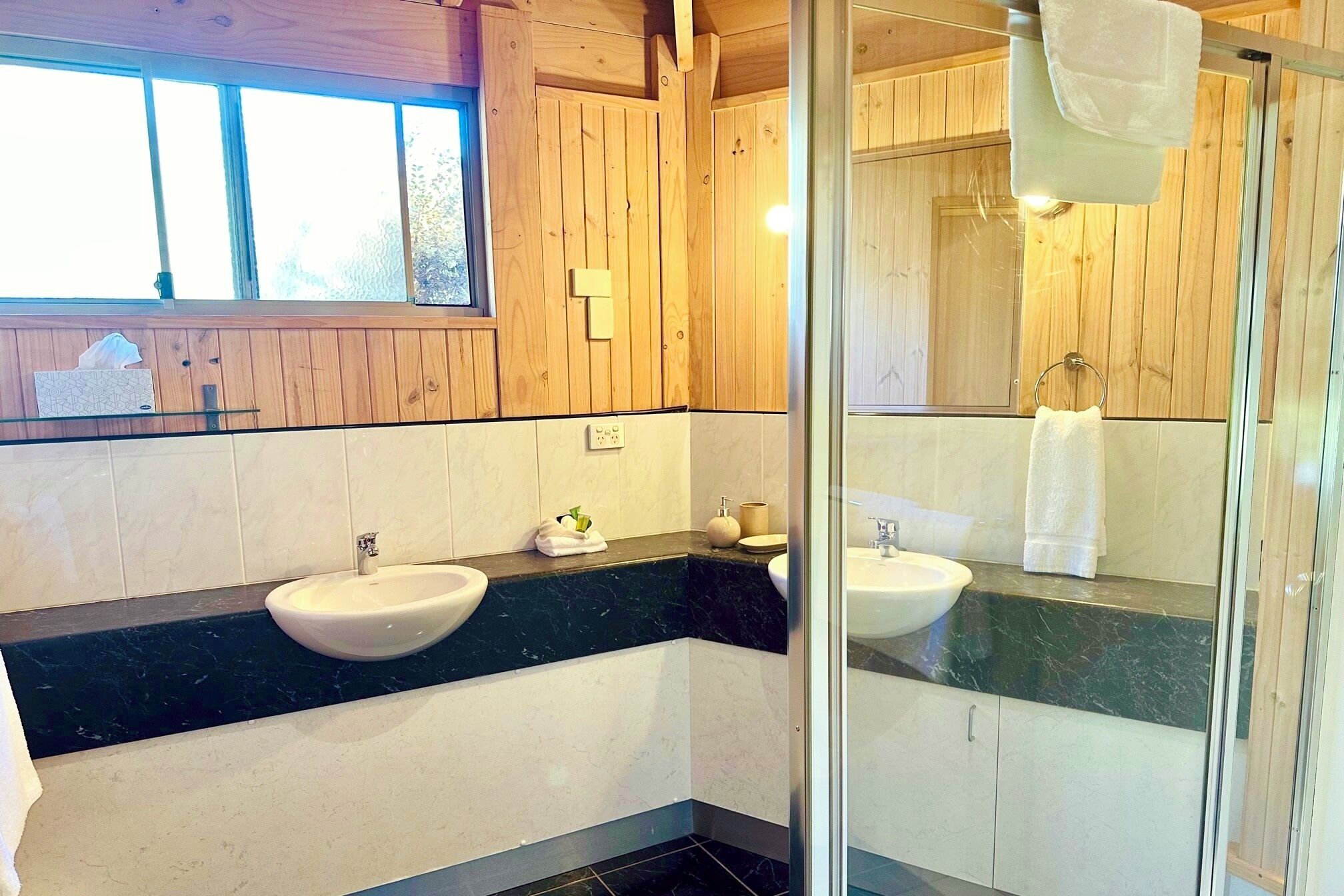 Un baño con paneles de madera, un lavamanos blanco, una encimera negra y una cabina de ducha de vidrio.