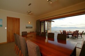 A spacious dining area with a large wooden table and chairs, a view of the ocean through an open door, and a ceiling with hanging lights.
