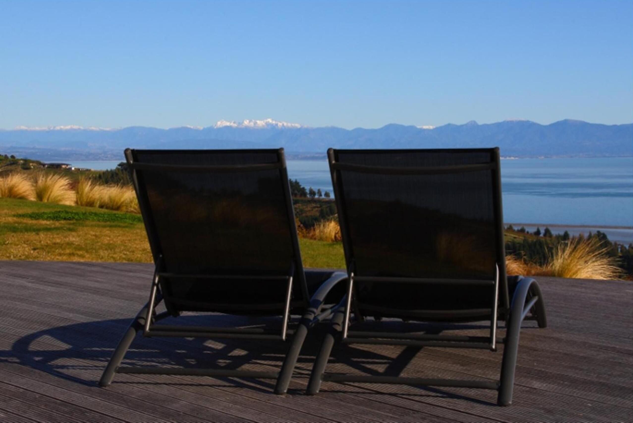 Two black lounge chairs on a wooden deck with a view of mountains and a lake.