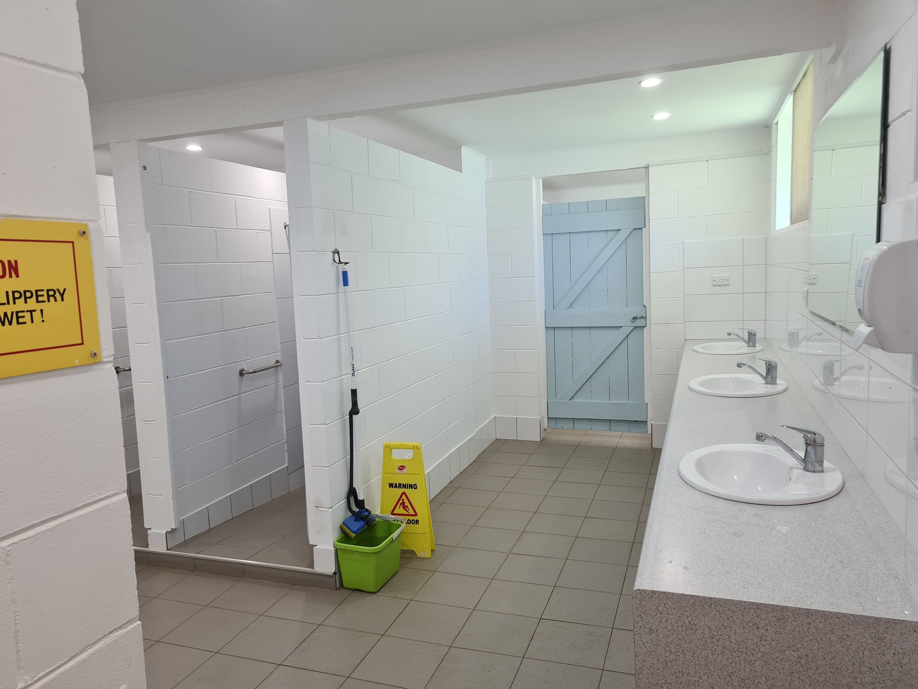 Un baño público con paredes de azulejos blancos, varios lavamanos y una zona de ducha.