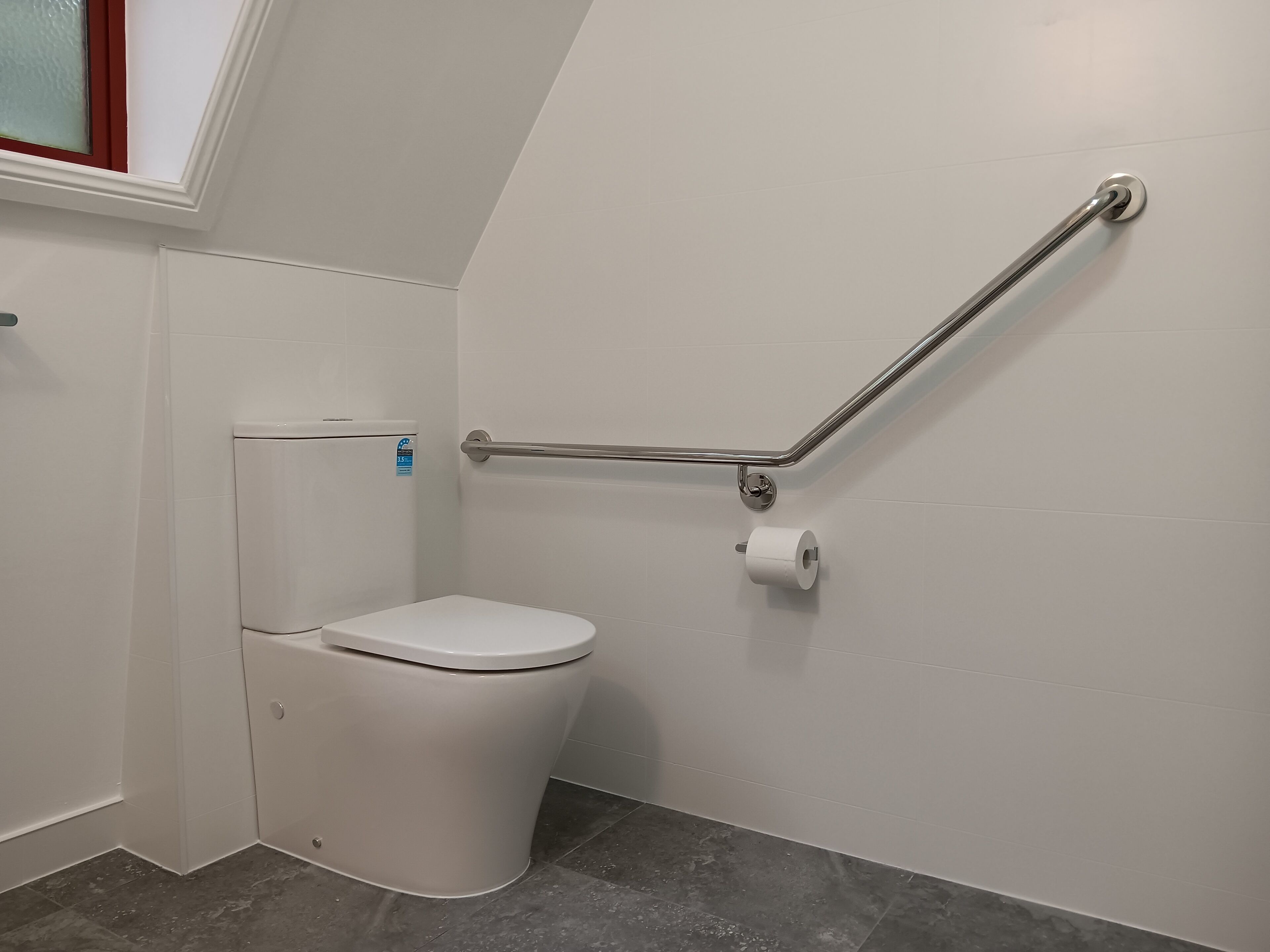 Sanitário branco com barra de apoio e rolo de papel higiénico na parede.
