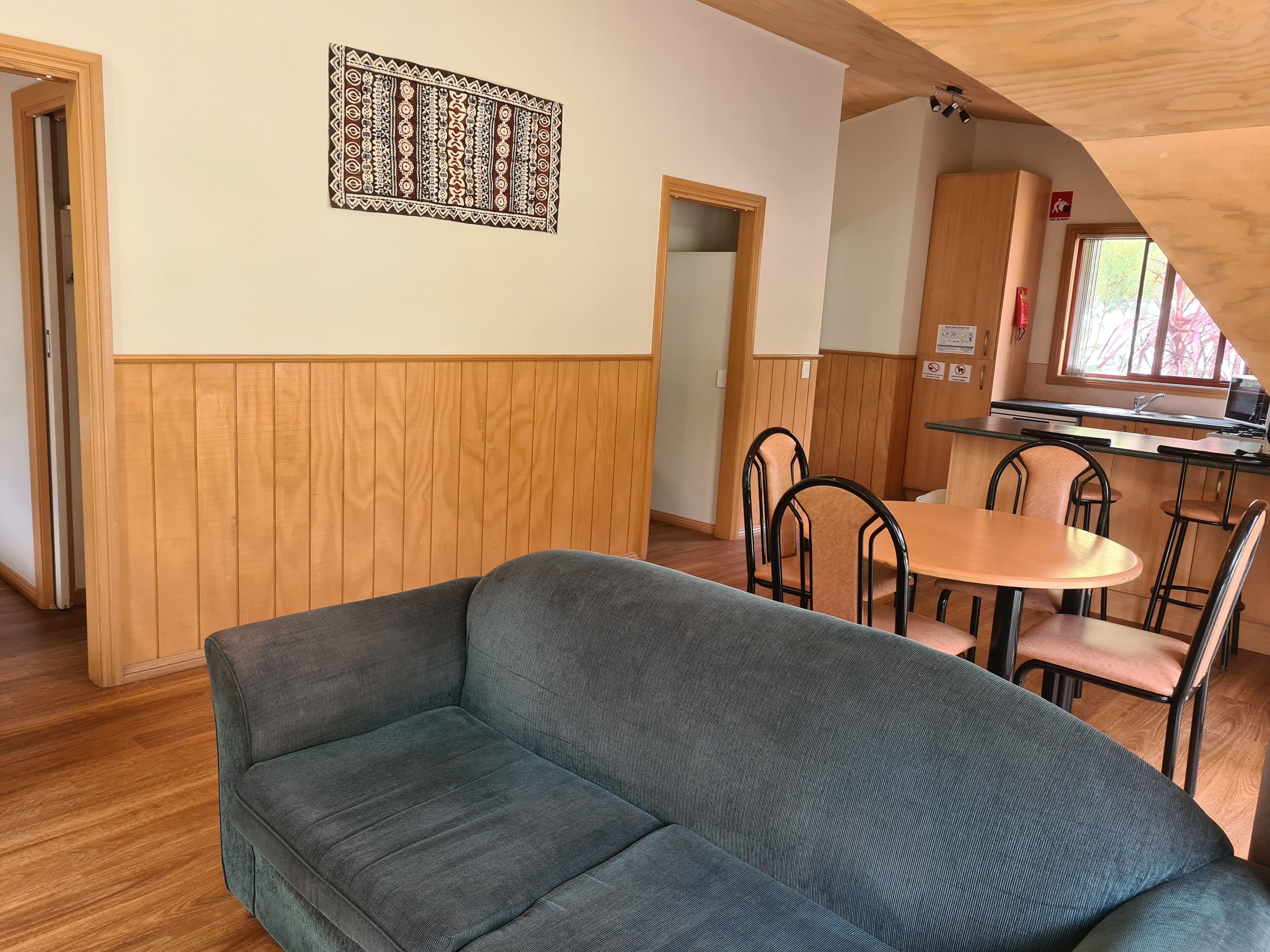 A cozy room with a grey sofa, wooden panel walls, a dining area with a table and chairs, and a kitchen visible through an open doorway.