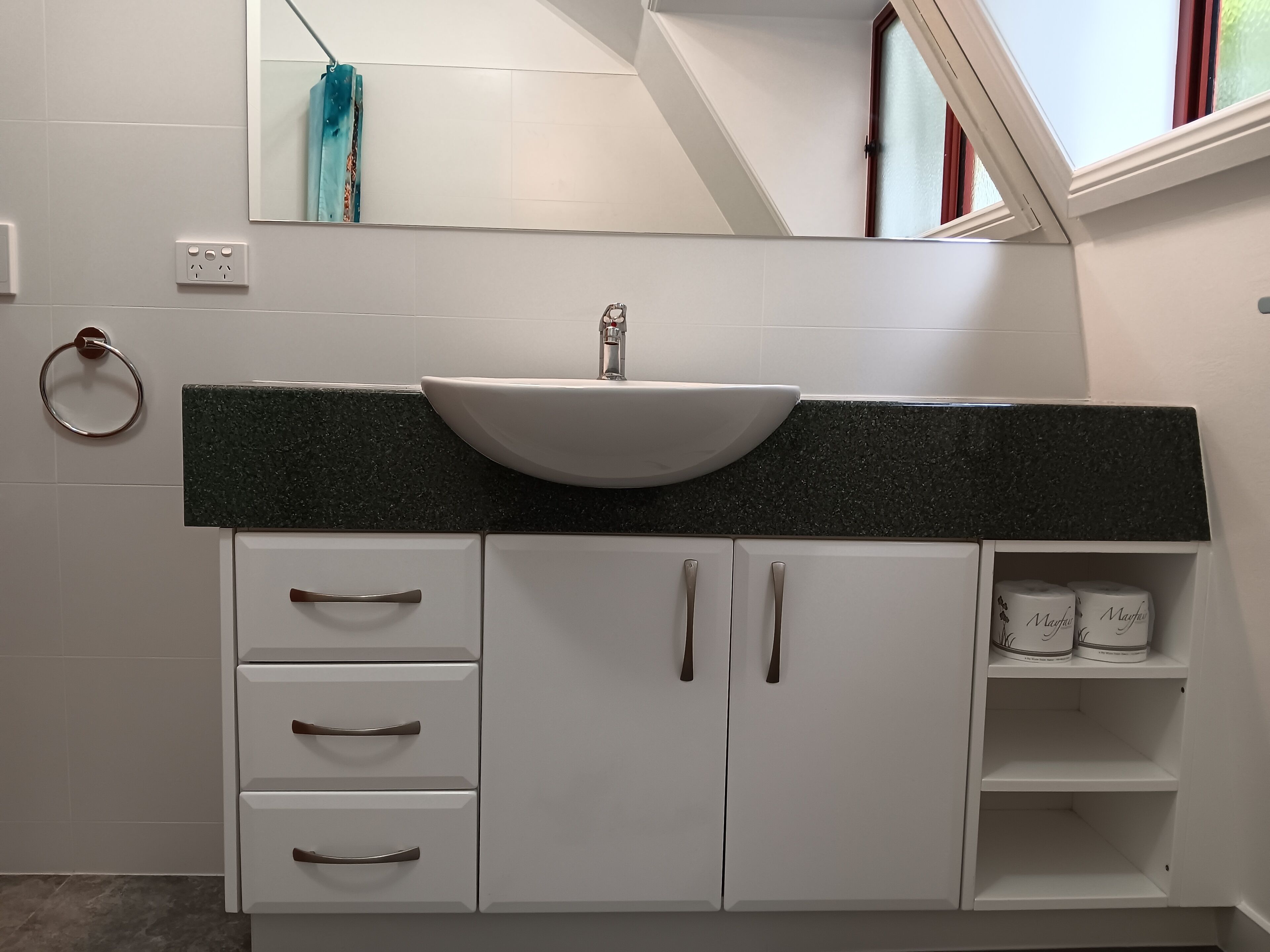 Une salle de bain avec un lavabo blanc, un plan de toilette en granit foncé, des armoires blanches avec des poignées argentées, un porte-serviettes et une étagère avec du papier toilette.