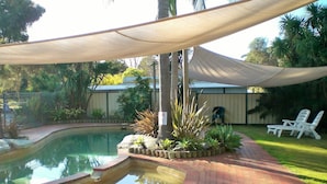 A swimming pool with a brick pathway, surrounded by plants and a shaded area with chairs.
