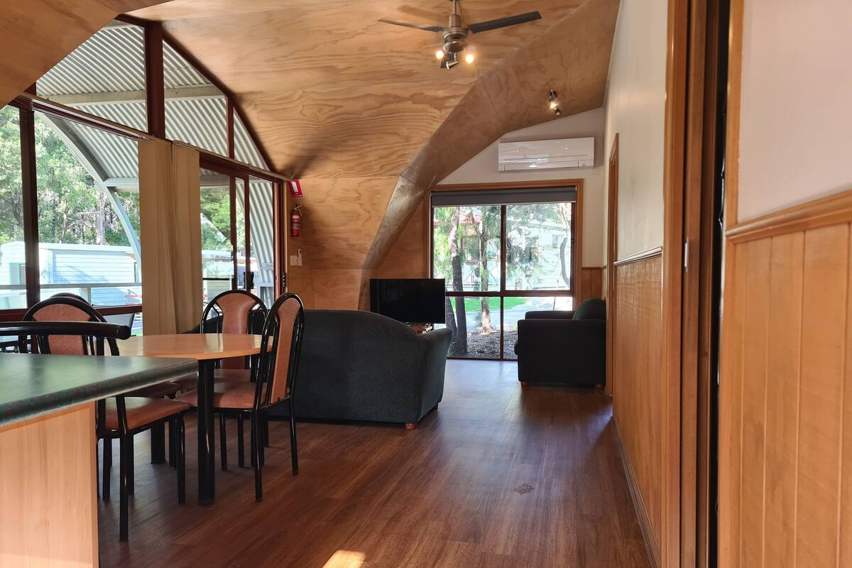 Un intérieur spacieux avec un plafond haut, un plancher en bois et de grandes fenêtres offrant une vue sur l’extérieur.