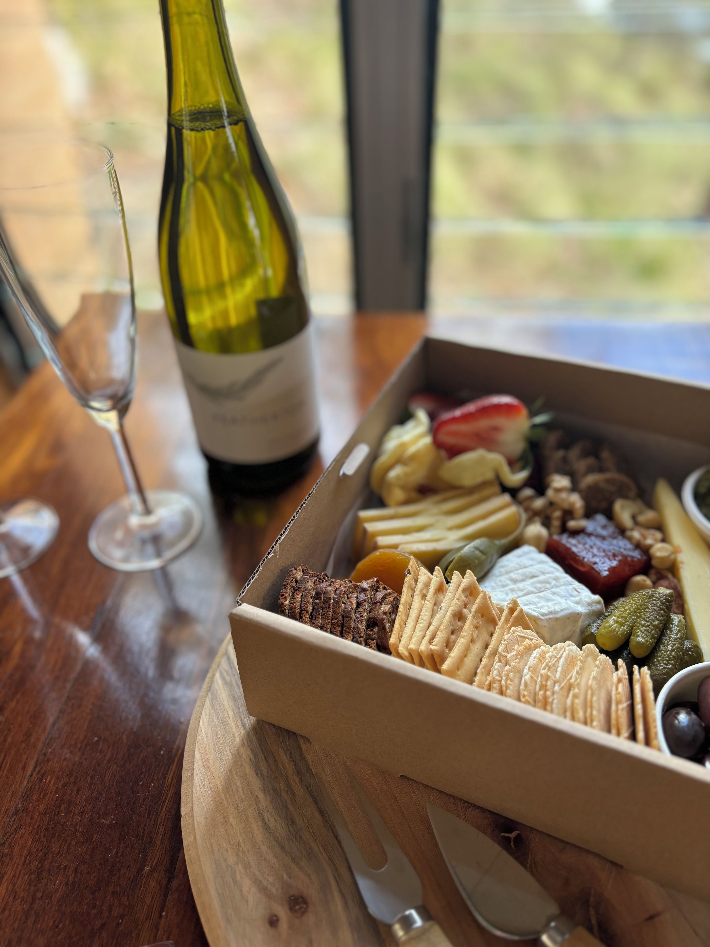 Una mesa de madera con una botella de vino, una copa y una tabla de fiambres con varios quesos, carnes y galletas.