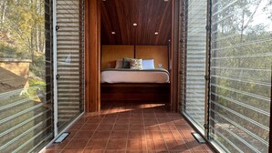 A bedroom with a wooden ceiling, a bed with pillows, and a view of trees through large windows. - The Odd Frog (Bright)