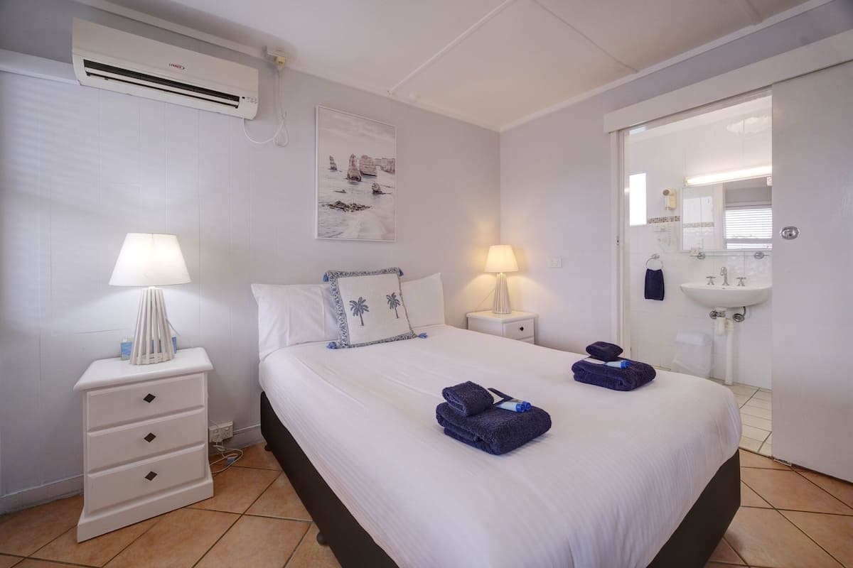 A hotel room with a bed, two bedside tables with lamps, a wall-mounted air conditioner, a painting, and a bathroom visible through an open door.