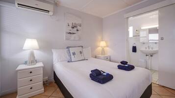 A hotel room with a bed, two bedside tables with lamps, a wall-mounted air conditioner, a painting, and a bathroom visible through an open door.