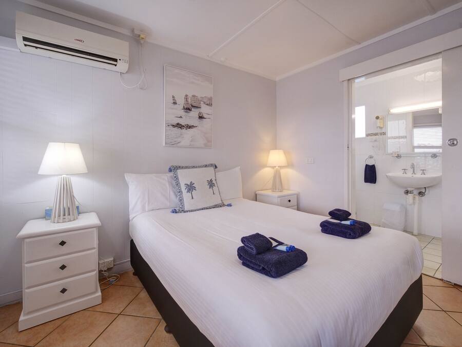 A hotel room with a bed, two bedside tables with lamps, a wall-mounted air conditioner, a painting, and a bathroom visible through an open door.
