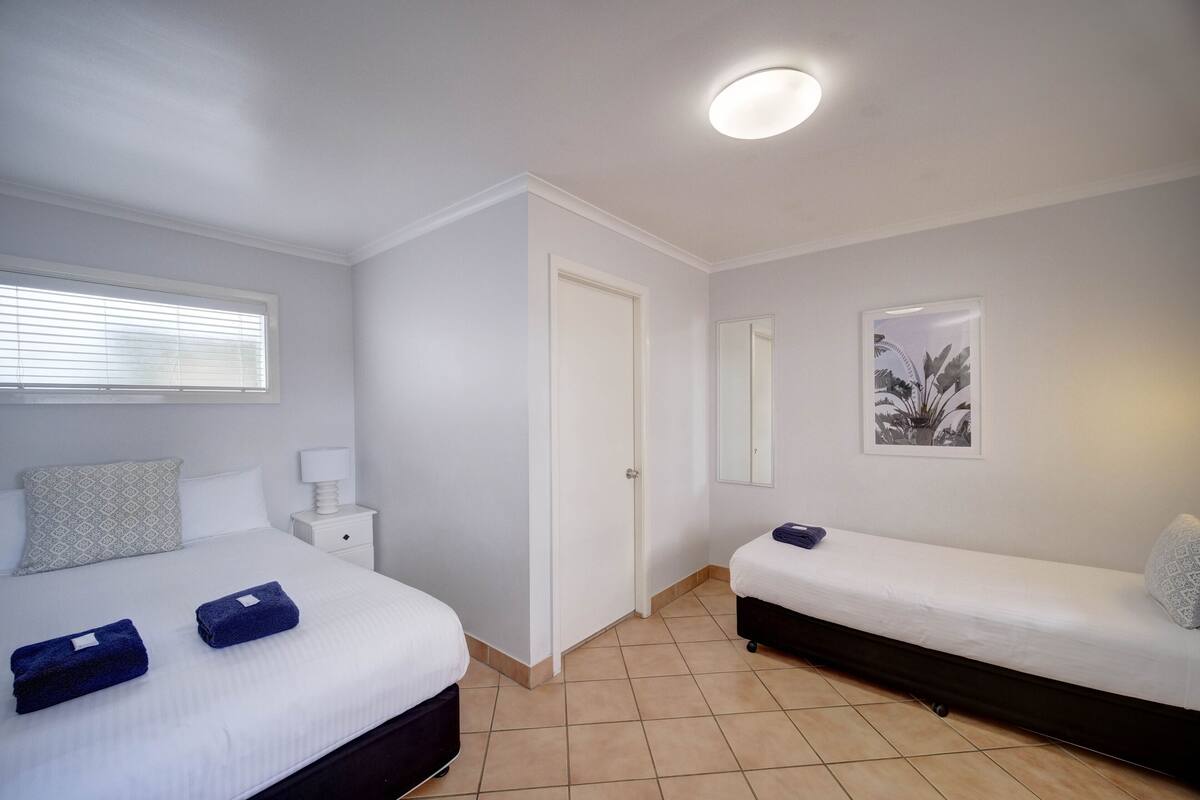 A hotel room with a bed, a bedside table, a nightstand, a window with blinds, a door, a wall-mounted mirror, and a framed picture.