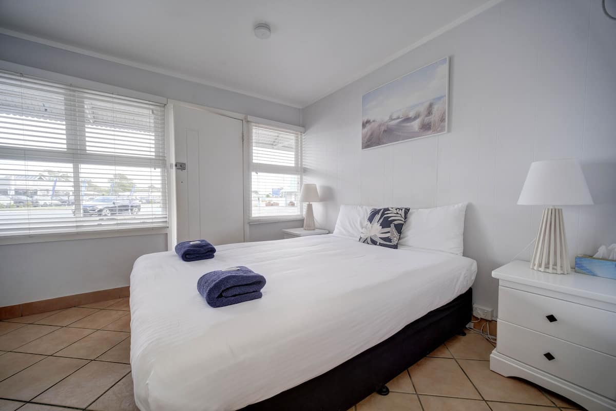 A hotel room with a large bed, white walls, a painting, and a window with blinds.
