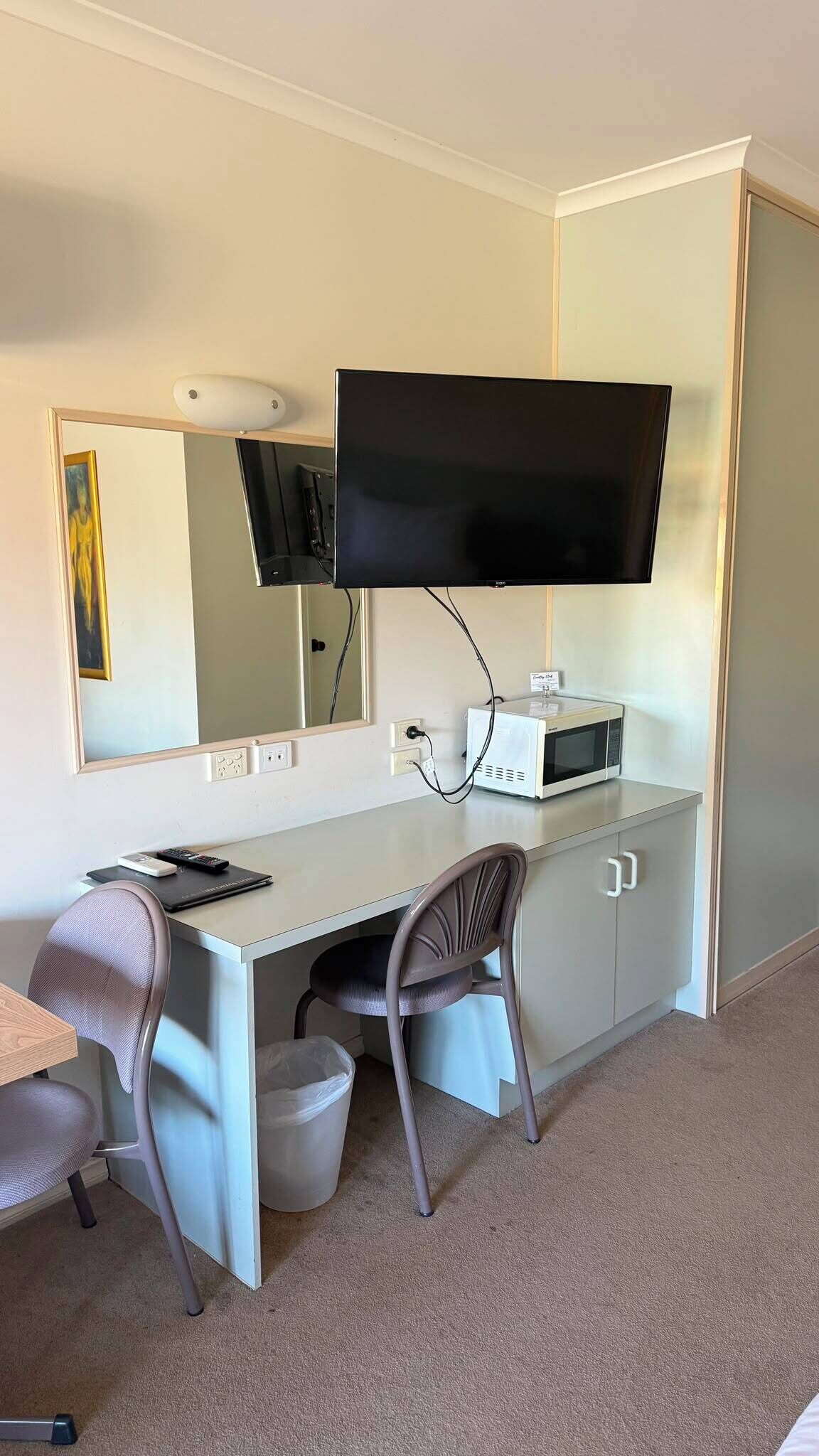 A hotel room with a desk, a chair, a television mounted on the wall, a microwave, and a trash bin.