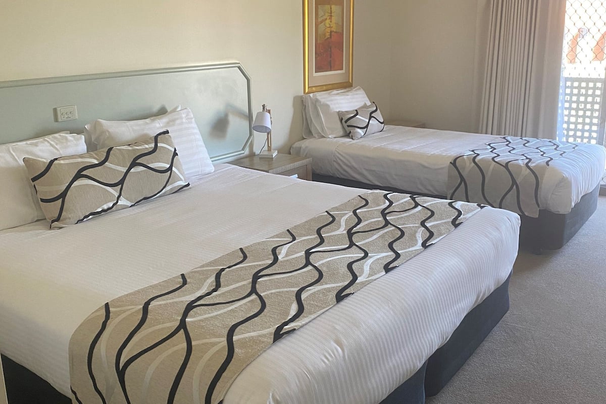 Ein Hotelzimmer mit zwei Betten, jeweils mit gemusterten Bettdecken und Kissen. An der Wand hängt ein Gemälde und es gibt ein Fenster mit Vorhängen.