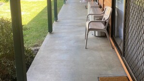 A covered outdoor seating area with tables and chairs, a concrete floor, and a view of a green lawn and picnic tables in the background.