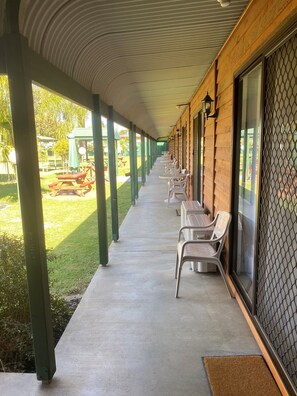 A covered outdoor seating area with tables and chairs, a concrete floor, and a view of a green lawn and picnic tables in the background. - Deniliquin Country Club Motor Inn (Deniliquin)