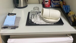 A kitchenette with a kettle, a coffee maker, a tea kettle, a microwave, a coffee cup, a glass, a tea bag, a packet of pringles, a packet of nobby's peanuts, and a packet of thins.