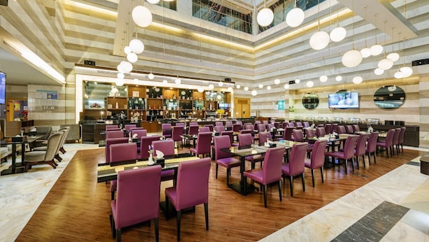 Un vaste espace dédié à la salle à manger, avec des chaises et des tables violettes, un coin bar équipé d’un téléviseur, et un plafond haut orné de luminaires suspendus.