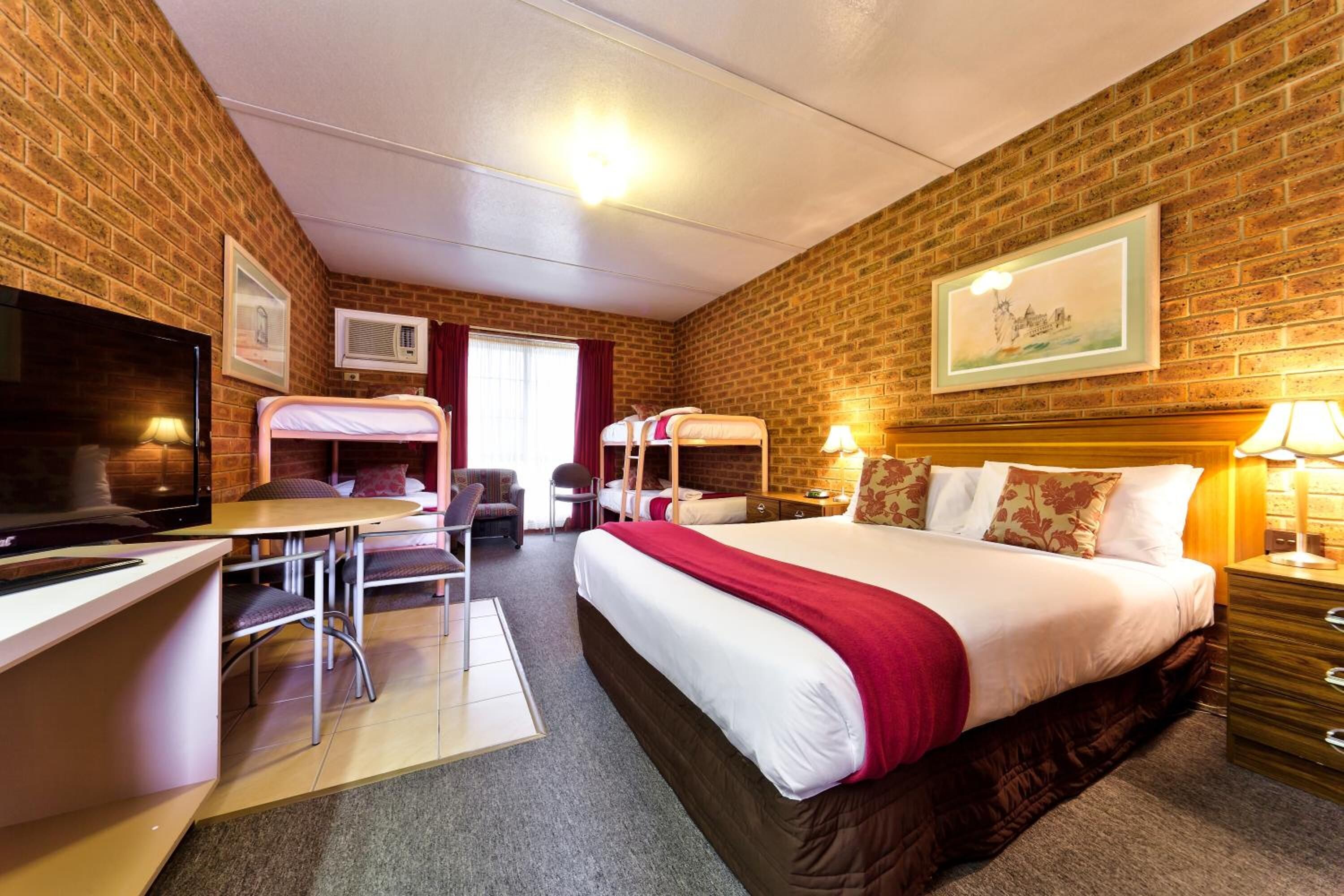 Habitación de hotel con pared de ladrillo, cama con manta roja, escritorio con silla, televisor y un cuadro en la pared.