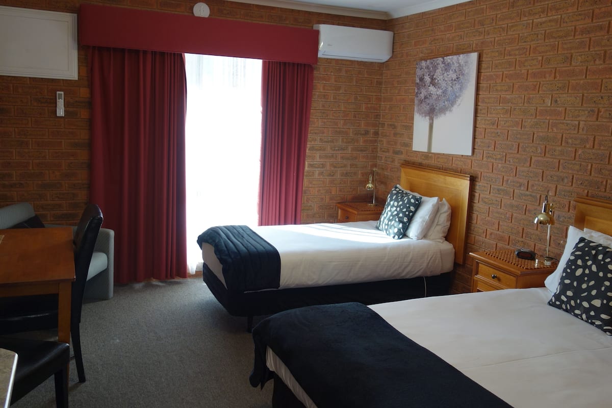 Una habitación de hotel con pared de ladrillo, cama con cabecera de madera, escritorio con silla y ventana con cortinas rojas.