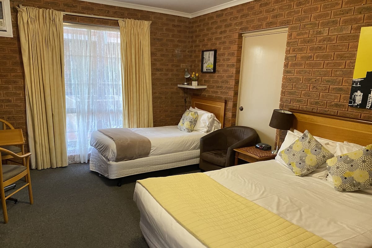 Una habitación de hotel con dos camas, una silla de madera, una mesita de noche y una pared de ladrillo.
