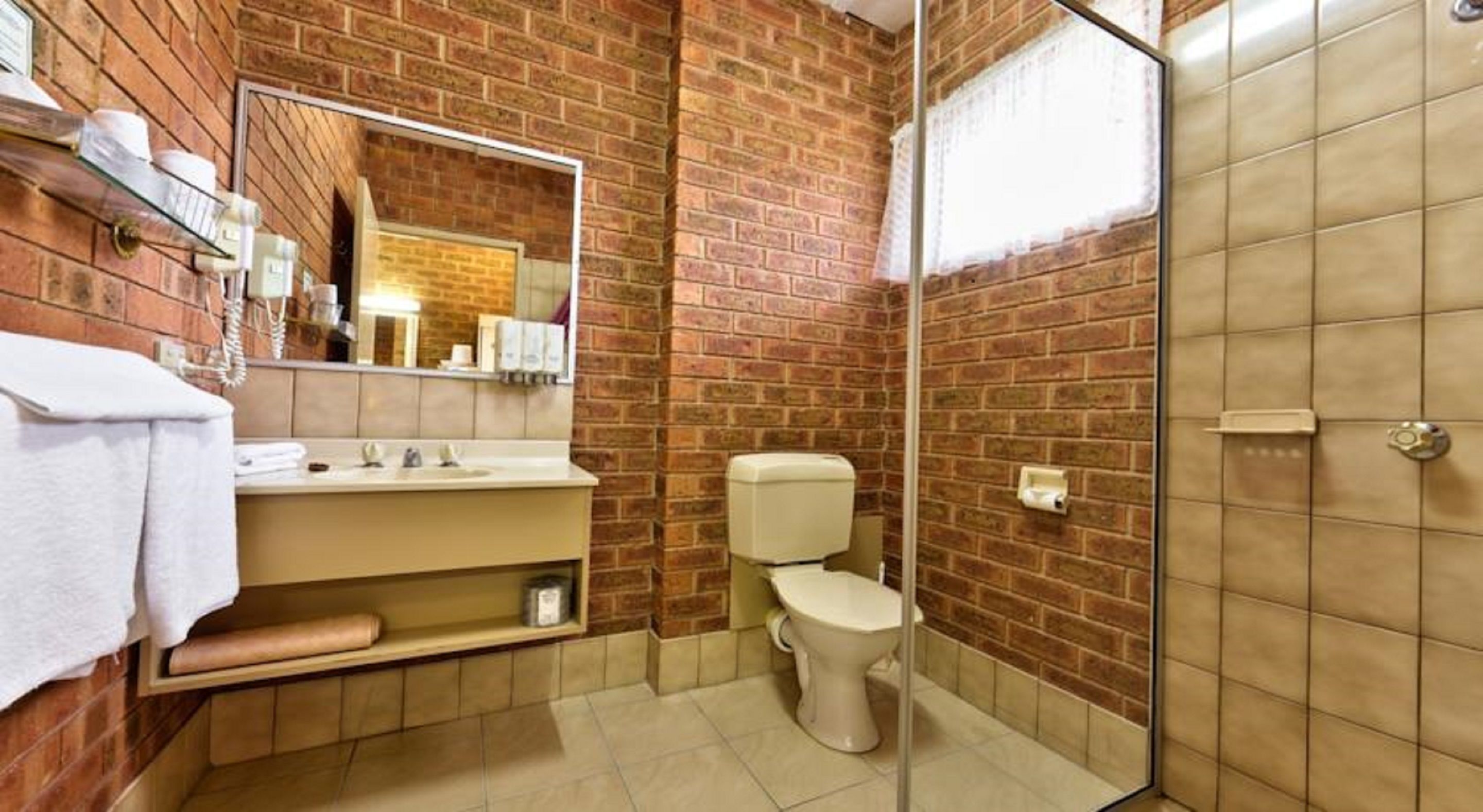 Baño con pared de ladrillo, cabina de ducha de vidrio, inodoro y lavamanos con espejo superior.