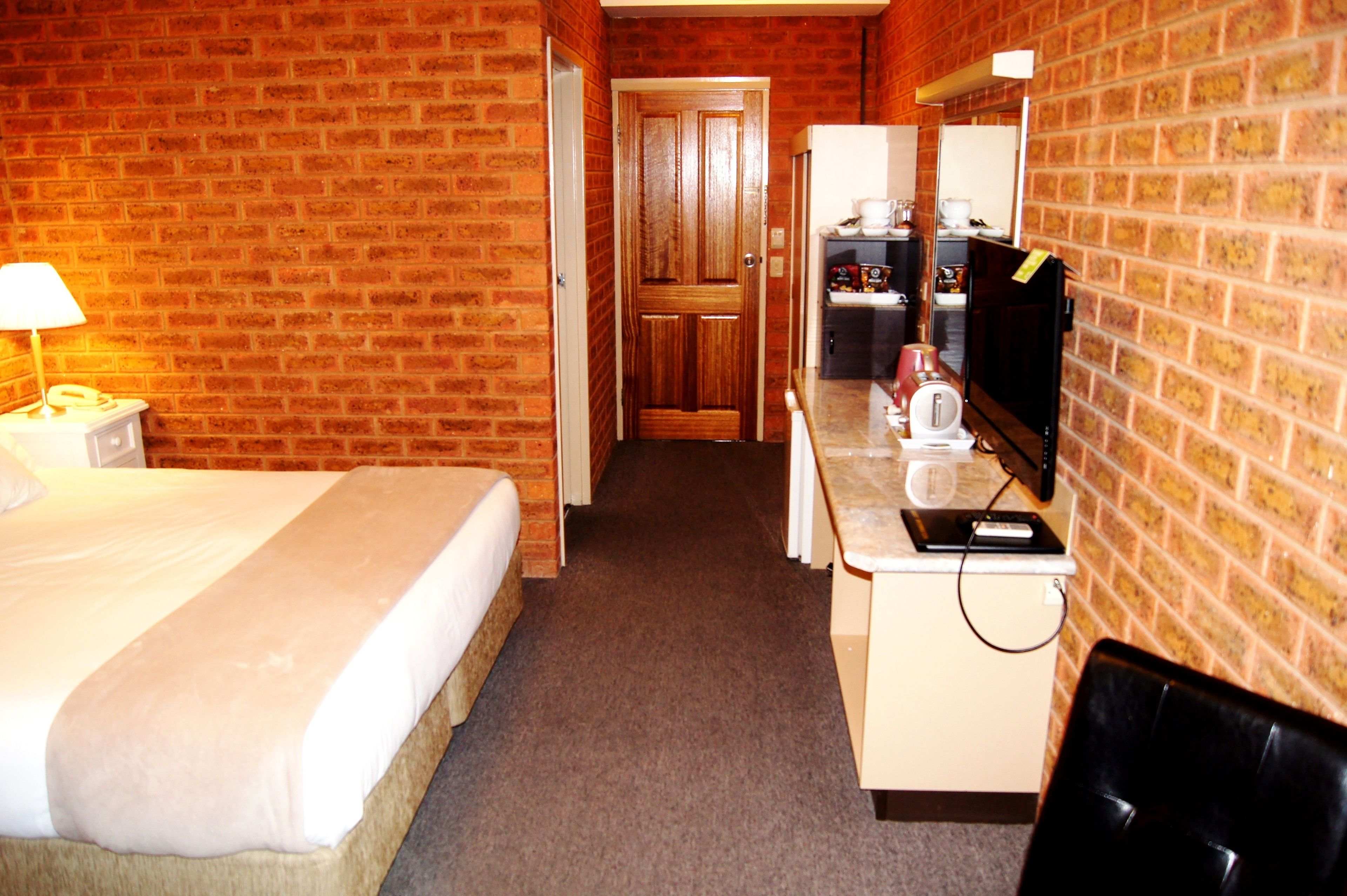 A hotel room with a brick wall, a bed with a white comforter, a desk with a computer and a flat-screen TV, and a small kitchenette.