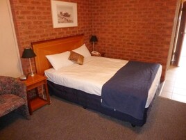 A hotel room with a brick wall, a wooden headboard, a bed with white linens, a blue blanket, and a small bedside table with a lamp.