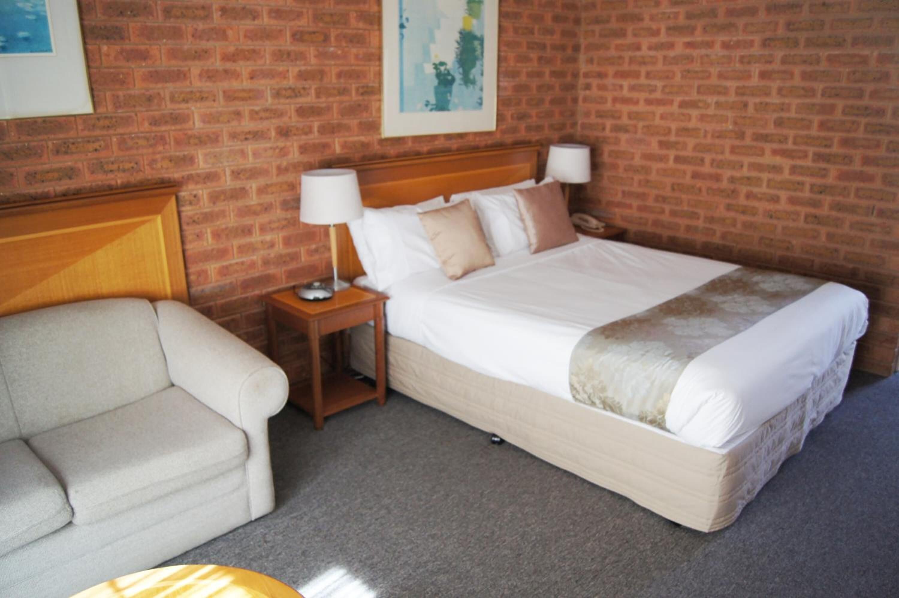 A hotel room with a brick wall, a bed with white bedding, a wooden headboard, a nightstand with a lamp, and a beige sofa.