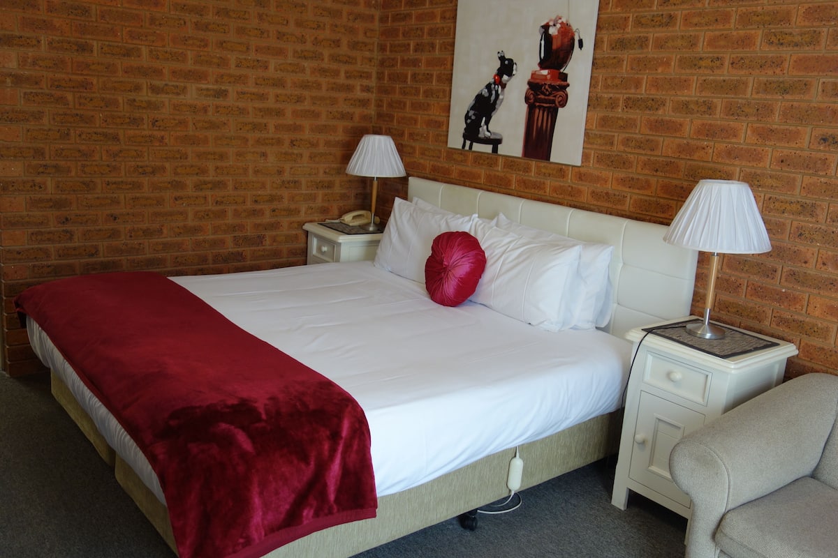 Una habitación de hotel con pared de ladrillo, una cama con ropa blanca, una manta roja, dos lámparas de noche, una mesita de noche con un teléfono y un cuadro en la pared.