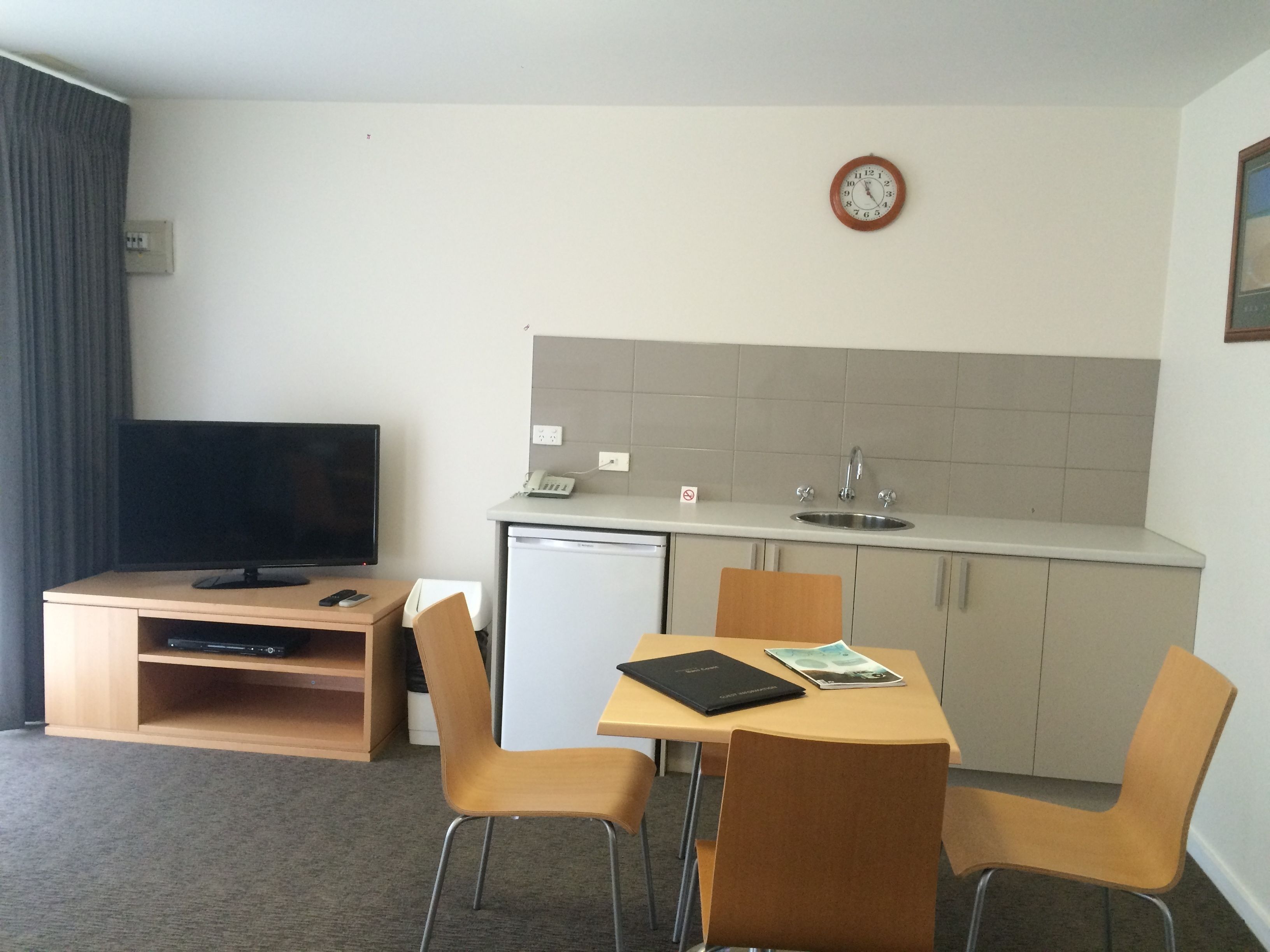 Een compacte hotelkamer met een kitchenette, een kleine koelkast, een flatscreen-tv, een eettafel met vier stoelen en een klok aan de muur.