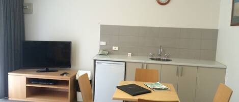 Habitación de hotel compacta con kitchenette, un pequeño refrigerador, televisor de pantalla plana, mesa de comedor con cuatro sillas y un reloj en la pared.