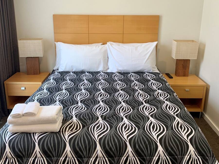A neatly made bed with a patterned bedspread, two white pillows, and a stack of folded towels on the foot of the bed. There are two bedside tables with lamps and a framed picture on the wall above the bed.