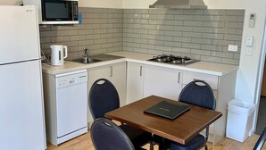Een compacte keuken met een magnetron, fornuis en vaatwasser. Er staat een houten tafel met vier stoelen en een kleine prullenbak.