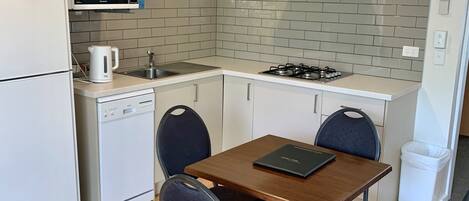 A compact kitchen with a microwave, stove, and dishwasher. There is a wooden table with four chairs and a small trash bin.