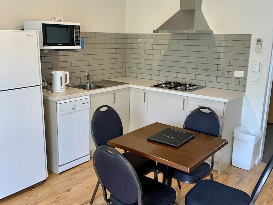 Una cocina compacta equipada con microondas, estufa y lavavajillas. Hay una mesa de madera con cuatro sillas y un pequeño cubo de basura.