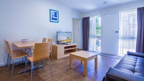 Een moderne woonkamer met een bank, een eettafel met stoelen, een televisie en een groot raam met gordijnen.