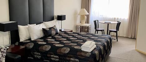 Habitación de hotel con una cama grande, un escritorio con silla, una lámpara y vistas al exterior a través de una ventana.