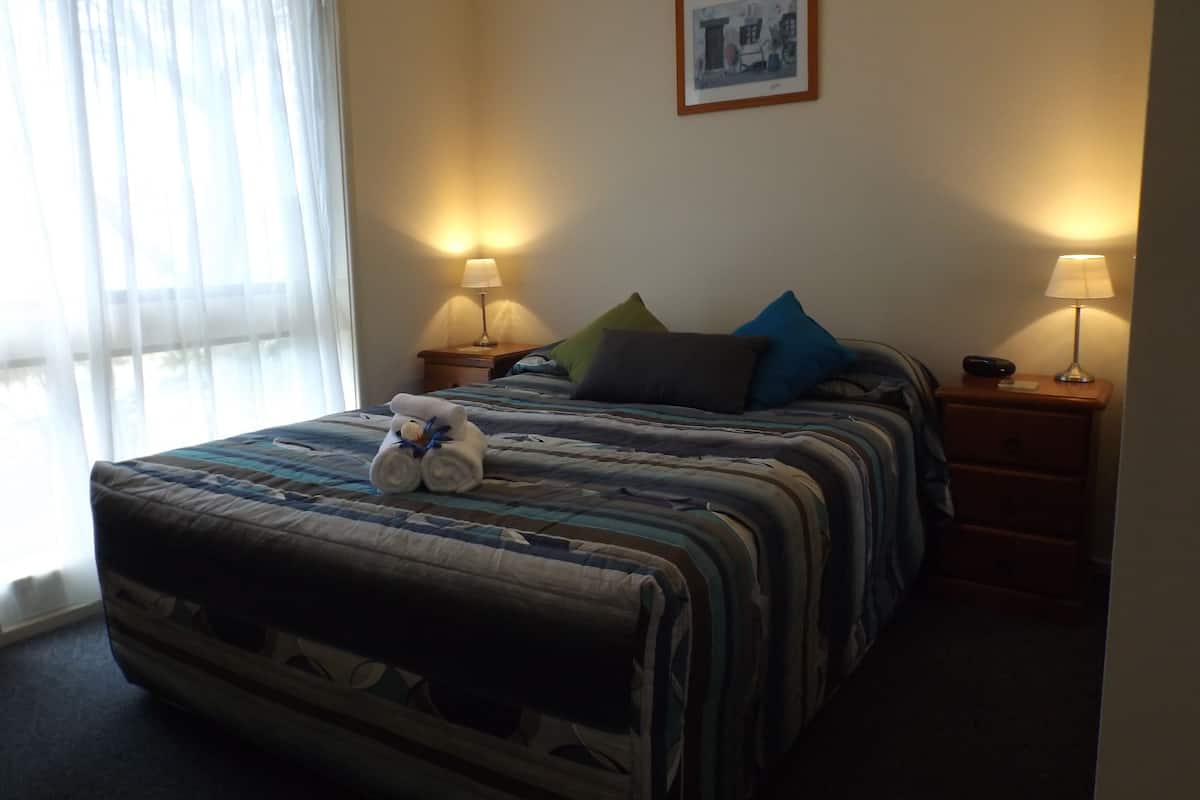 Habitación de hotel con cama, dos mesitas de noche con lámparas, un cuadro en la pared y una ventana con cortinas.