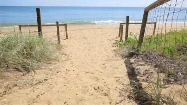 Un sentier de sable qui longe la plage et mène à l’océan, bordé de poteaux en bois et d’une clôture en fil de fer.
