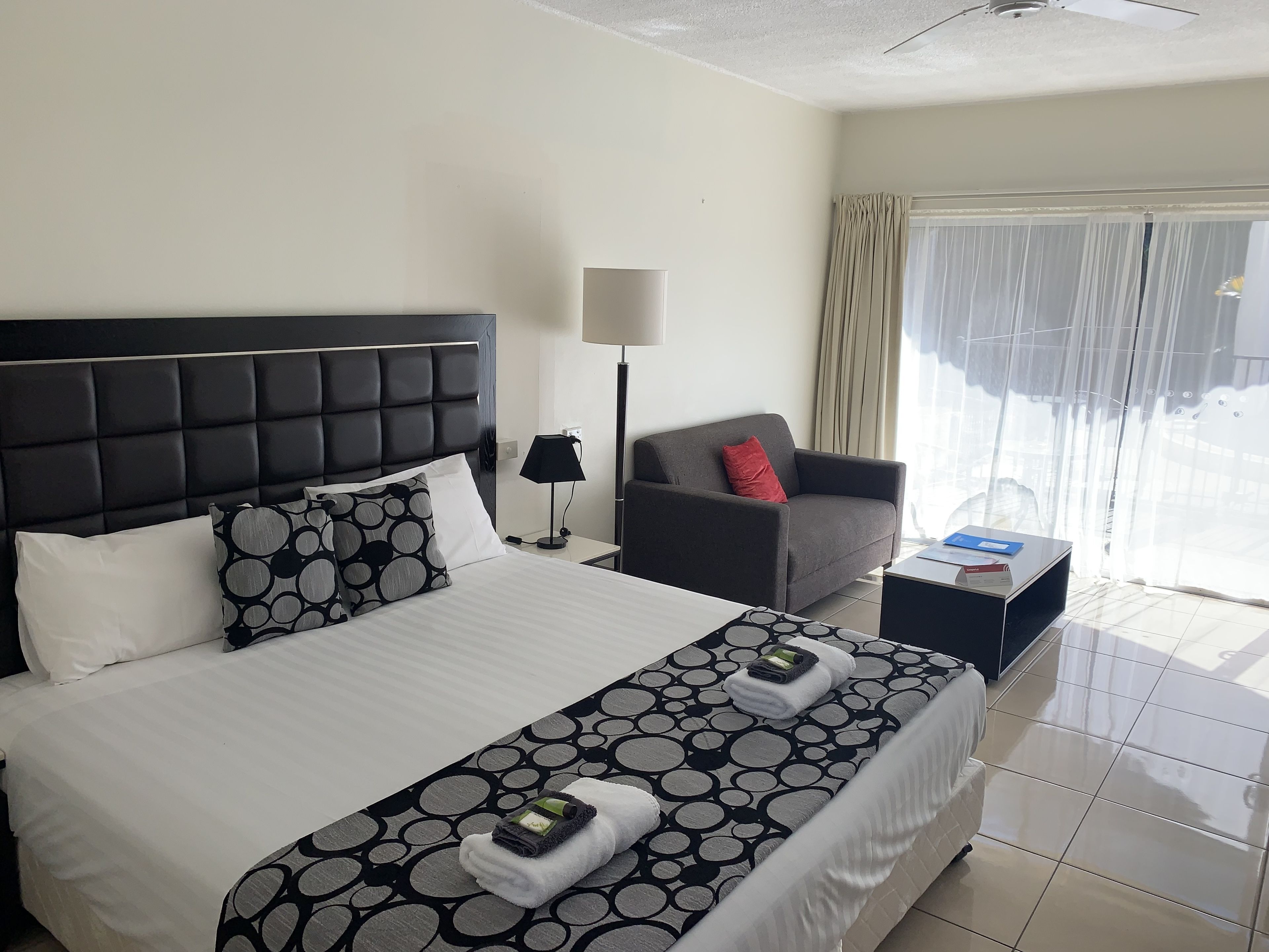 Habitación de hotel con una cama grande, un sillón gris, una mesita de noche con lámpara y vistas al exterior a través de una ventana con cortinas.