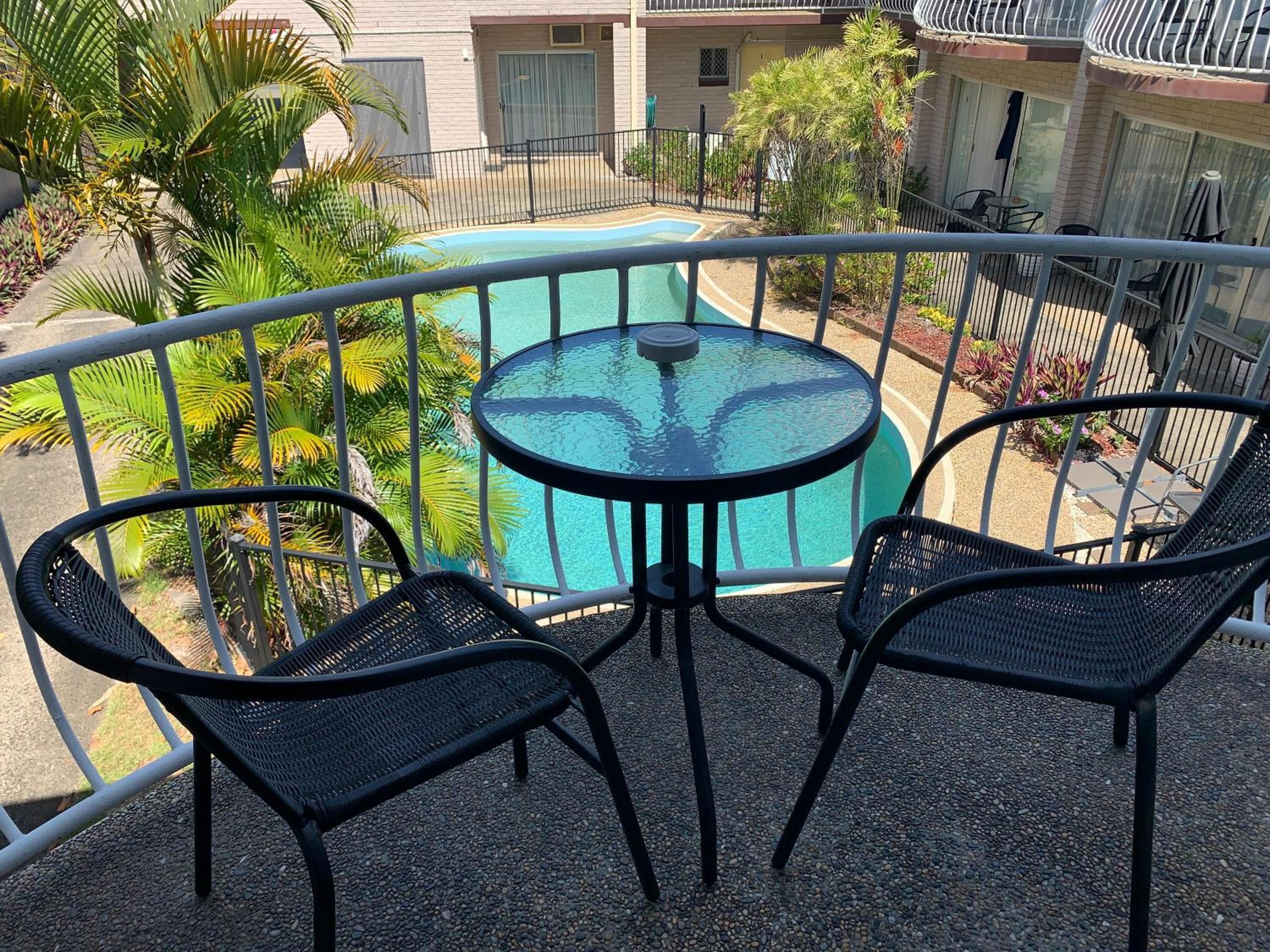 A balcony with a glass table and two chairs, overlooking a swimming pool and a building with a balcony.